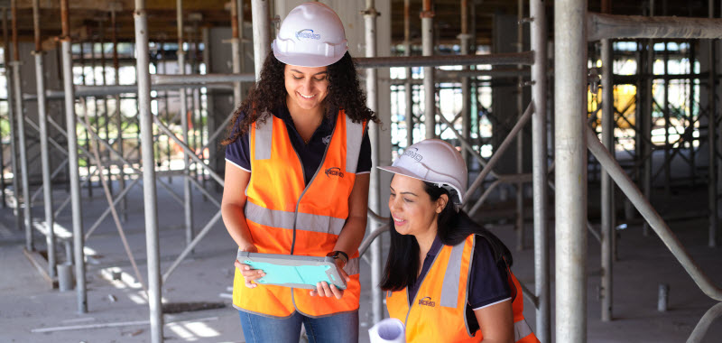 Two women wearing safety gears looking at a tablet while in the construction site