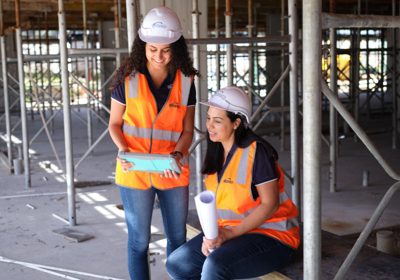 Two women wearing safety gears looking at a tablet while in the construction site